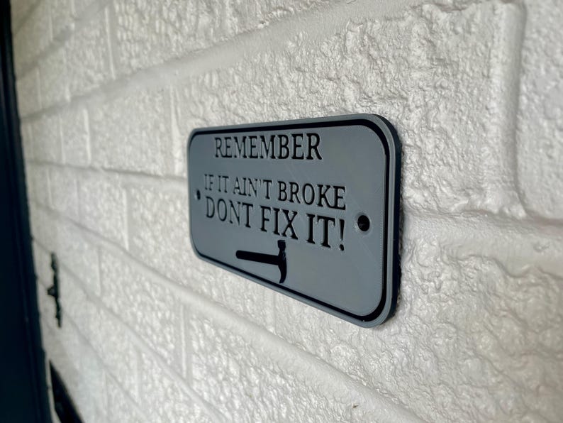 May include: A rectangular gray metal sign with black text that reads "REMEMBER IF IT AIN'T BROKE DON'T FIX IT!" A black hammer graphic is at the bottom. The sign is mounted on a white brick wall.