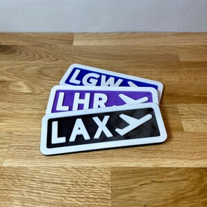 May include: Three airport code signs with white lettering and a white airplane icon on a black, purple, and blue background. The signs read "LAX", "LHR", and "LGW".