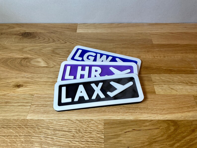 May include: Three airport code signs with white lettering and a white airplane icon on a black, purple, and blue background. The signs read "LAX", "LHR", and "LGW".