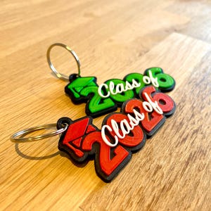 May include: Two graduation-themed keychains. One is green, the other red, both with the text "Class of 2026" in white. Each keychain has a silver ring attached. The keychains are on a wooden surface.