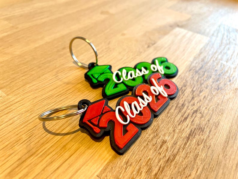 May include: Two graduation-themed keychains. One is green, the other red, both with the text "Class of 2026" in white. Each keychain has a silver ring attached. The keychains are on a wooden surface.