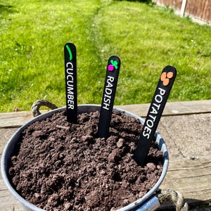 May include: Three black plant markers with white text and colorful icons. The markers are labeled "Cucumber", "Radish", and "Potatoes".
