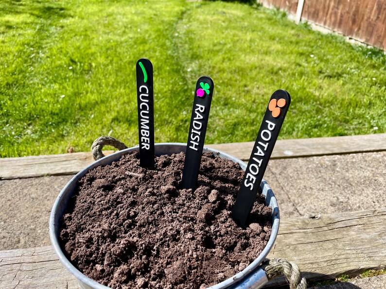 May include: Three black plant markers with white text and colorful icons. The markers are labeled "Cucumber", "Radish", and "Potatoes".