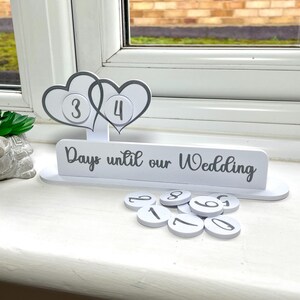 May include: White wooden countdown calendar with two hearts and the text "Days until our Wedding". The calendar has a number of white circles with black numbers on them.