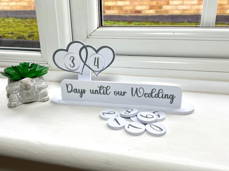 May include: White wooden countdown calendar with two hearts and the text "Days until our Wedding". The calendar has a number of white circles with black numbers on them.