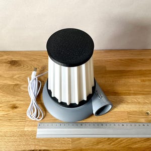 Puede incluir: Un dispositivo impreso en 3D blanco y gris con una parte superior negra y un cable USB. El dispositivo tiene una base redonda y una parte superior cil&iacute;ndrica con un dise&ntilde;o acanalado. El dispositivo tiene un peque&ntilde;o tubo gris que se extiende desde el lateral.