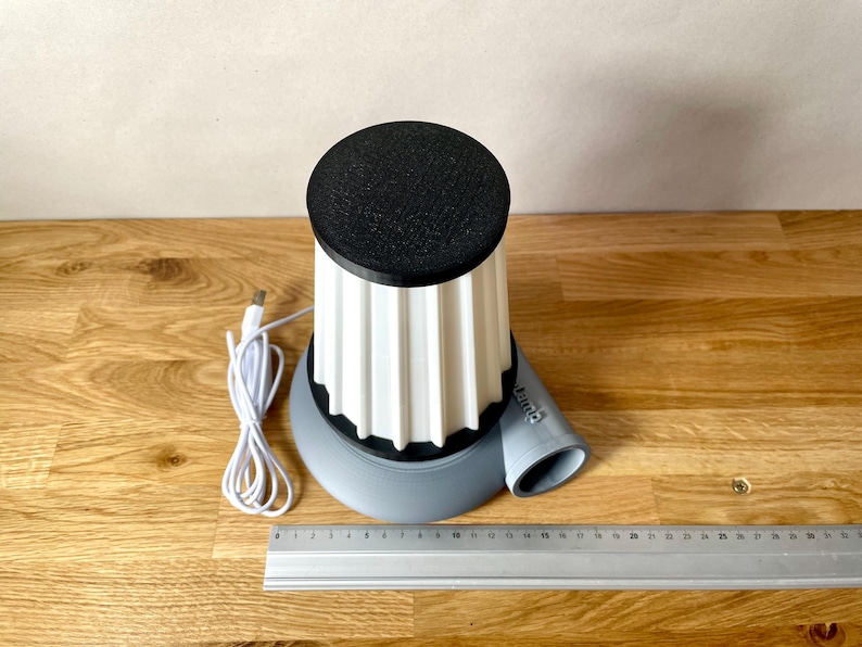 Puede incluir: Un dispositivo impreso en 3D blanco y gris con una parte superior negra y un cable USB. El dispositivo tiene una base redonda y una parte superior cil&iacute;ndrica con un dise&ntilde;o acanalado. El dispositivo tiene un peque&ntilde;o tubo gris que se extiende desde el lateral.
