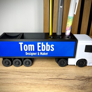 Pu&ograve; includere: Organizzatore da scrivania nero e bianco a forma di camion. Il pannello laterale blu mostra il testo "Tom Ebbs Designer & Maker" in bianco. Contiene penne, matite e altri articoli per l'ufficio.