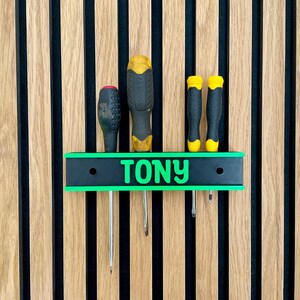 May include: A personalized screwdriver holder with the name "TONY" in green lettering. The holder is black with a green border and holds four screwdrivers with black and yellow handles. The background is a wooden wall with vertical stripes.