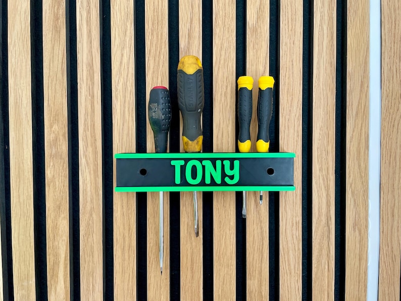 May include: A personalized screwdriver holder with the name "TONY" in green lettering. The holder is black with a green border and holds four screwdrivers with black and yellow handles. The background is a wooden wall with vertical stripes.