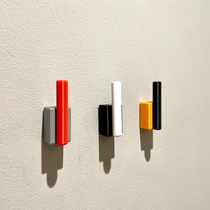 Op de afbeelding: Drie wandhaken met verschillende gekleurde accenten. De haak links heeft een grijze basis en een rood accent. De middelste haak heeft een zwarte basis en een wit accent. De haak rechts heeft een gele basis en een zwart accent.