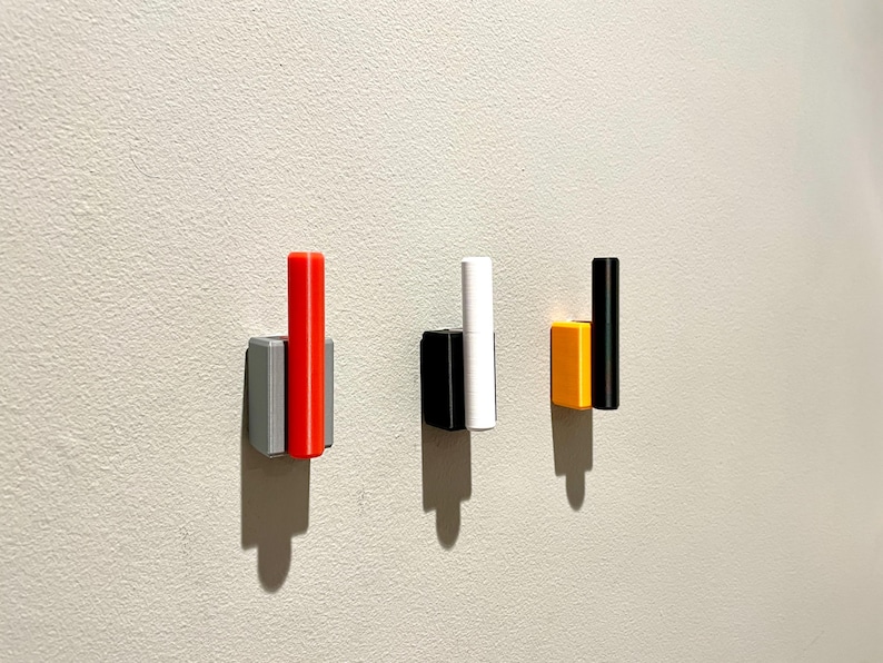 Op de afbeelding: Drie wandhaken met verschillende gekleurde accenten. De haak links heeft een grijze basis en een rood accent. De middelste haak heeft een zwarte basis en een wit accent. De haak rechts heeft een gele basis en een zwart accent.