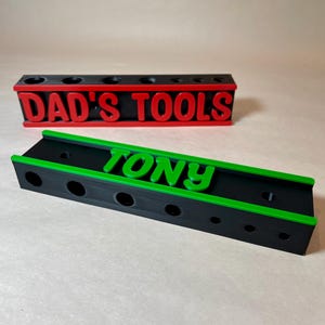 May include: Two tool organizers, one with "DAD'S TOOLS" in red letters and the other with "TONY" in green letters. Both are black with colored accents and circular holes for tool storage. The organizers are on a neutral surface.