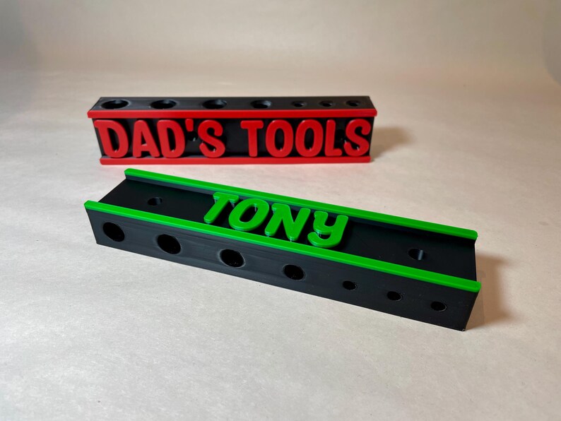 May include: Two tool organizers, one with "DAD'S TOOLS" in red letters and the other with "TONY" in green letters. Both are black with colored accents and circular holes for tool storage. The organizers are on a neutral surface.