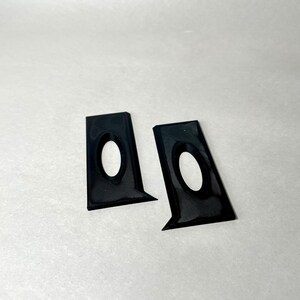 May include: Two black, angular brackets with an oval cutout. The brackets have a textured surface and a glossy finish. They are positioned on a white background, suggesting a product shot.
