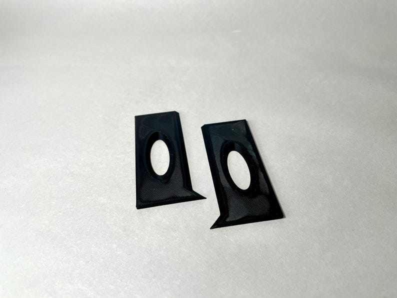 May include: Two black, angular brackets with an oval cutout. The brackets have a textured surface and a glossy finish. They are positioned on a white background, suggesting a product shot.