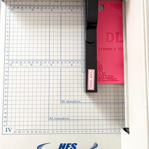 May include: A white paper cutter with a grid-marked base and a black cutting arm labeled TOOL BOX. A bright pink rectangular card with the text DL and 210mm x 99mm is positioned under the blade. The base features the HFS logo and website.