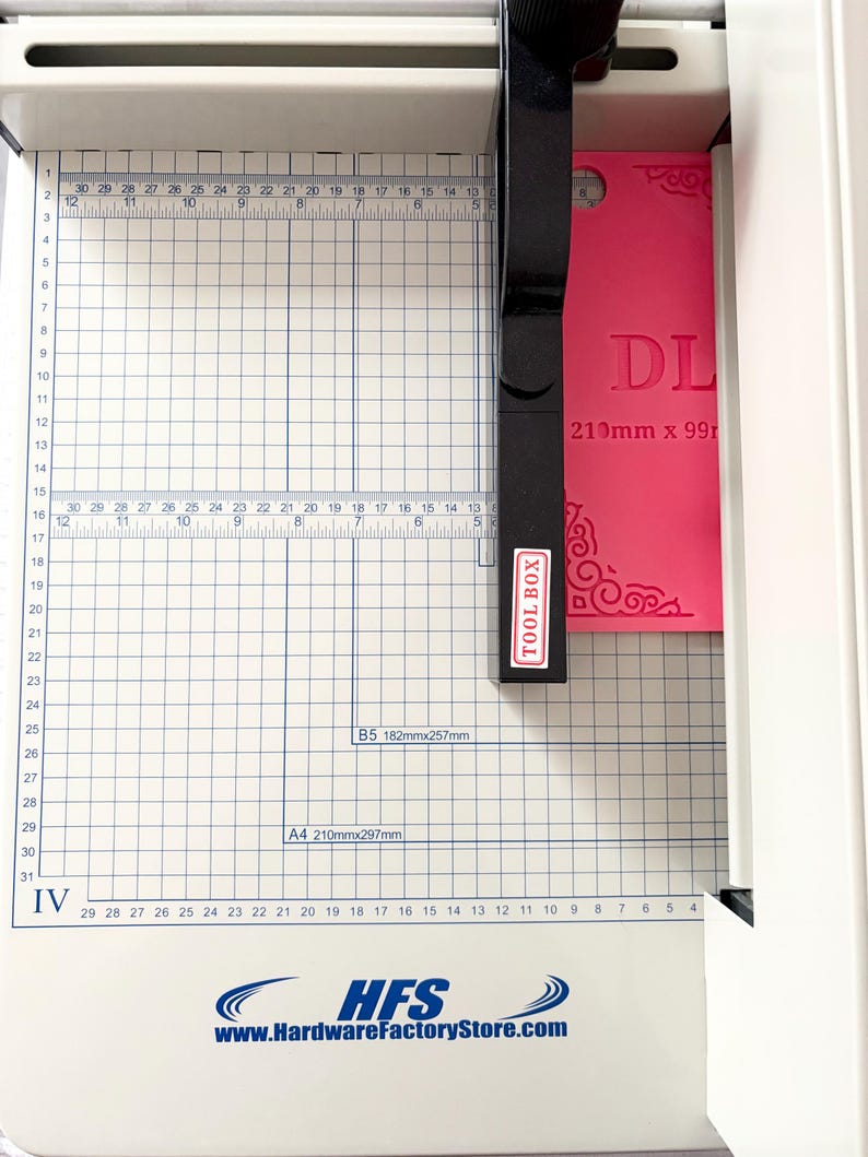 May include: A white paper cutter with a grid-marked base and a black cutting arm labeled TOOL BOX. A bright pink rectangular card with the text DL and 210mm x 99mm is positioned under the blade. The base features the HFS logo and website.