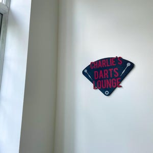 Triangular darts sign with the text charlie's darts lounge in red. Main background is black and has silver inlayed darts logos. On a white wall