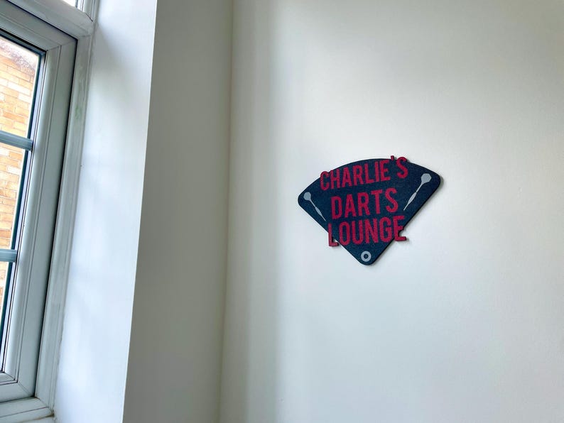 Triangular darts sign with the text charlie's darts lounge in red. Main background is black and has silver inlayed darts logos. On a white wall