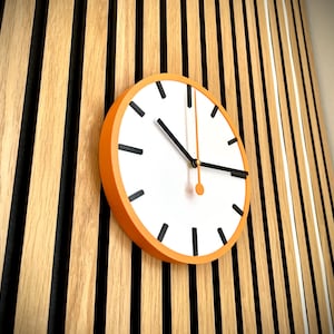May include: A white clock face with black hour and minute hands and black markings for the numbers. The clock has an orange frame and is mounted on a wall with vertical wooden slats.
