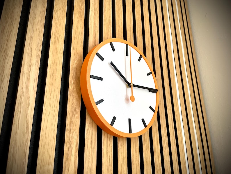 May include: A white clock face with black hour and minute hands and black markings for the numbers. The clock has an orange frame and is mounted on a wall with vertical wooden slats.