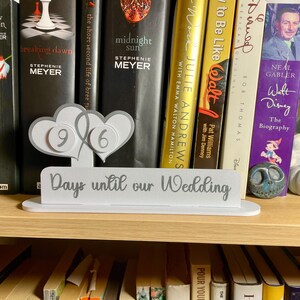 May include: A white wooden sign with the text "Days until our Wedding" in gray lettering. Two white hearts with gray numbers 9 and 6 are attached to the sign. The sign is sitting on a bookshelf with a variety of books.