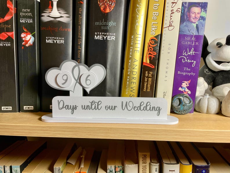 May include: A white wooden sign with the text "Days until our Wedding" in gray lettering. Two white hearts with gray numbers 9 and 6 are attached to the sign. The sign is sitting on a bookshelf with a variety of books.