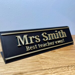 name plaque / plate with a teachers name and best teacher ever! text on the front. Black with bronze text on a wooden desk with grey background