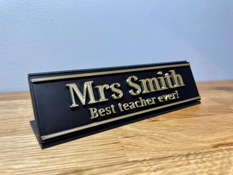 name plaque / plate with a teachers name and best teacher ever! text on the front. Black with bronze text on a wooden desk with grey background