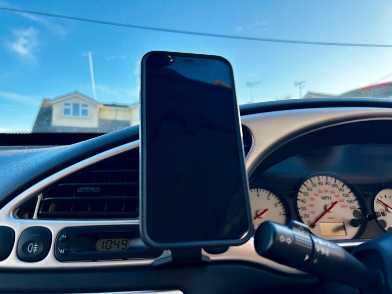 May include: A black smartphone is mounted in a car's dashboard using a phone holder. The phone's screen is black and the dashboard has a digital clock displaying 10:49.