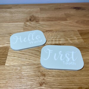 May include: Two white rectangular silicone stamps with raised lettering. The first stamp says "Hello World." The second stamp says "First Tooth."