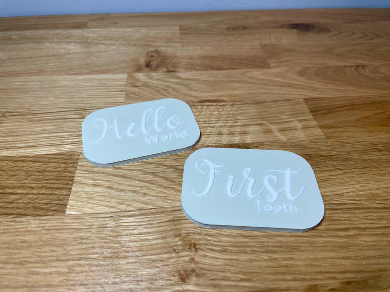 May include: Two white rectangular silicone stamps with raised lettering. The first stamp says "Hello World." The second stamp says "First Tooth."