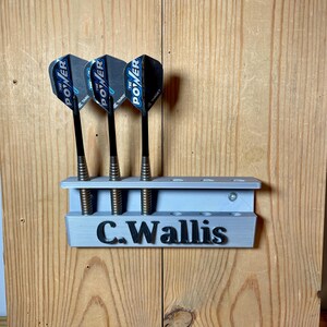 Silver / grey wall mounted dart holder with large black text along the lower edge showing a name. 3 darts in the holder with a total of 6 available slots. Shown mounted to a wooden background.