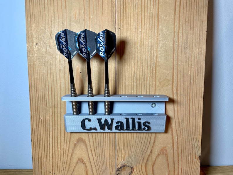 Silver / grey wall mounted dart holder with large black text along the lower edge showing a name. 3 darts in the holder with a total of 6 available slots. Shown mounted to a wooden background.