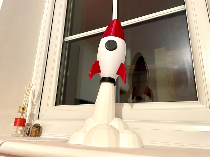 Op de afbeelding: Een witte en rode raketlamp met rode vinnen en een zwart cirkelvormig detail. De raket staat op een witte wolkvormige basis. De lamp staat op een vensterbank.