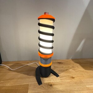 Op de afbeelding: Een zwarte en oranje tafellamp in de vorm van een schokdemper met een witte gloeilamp.