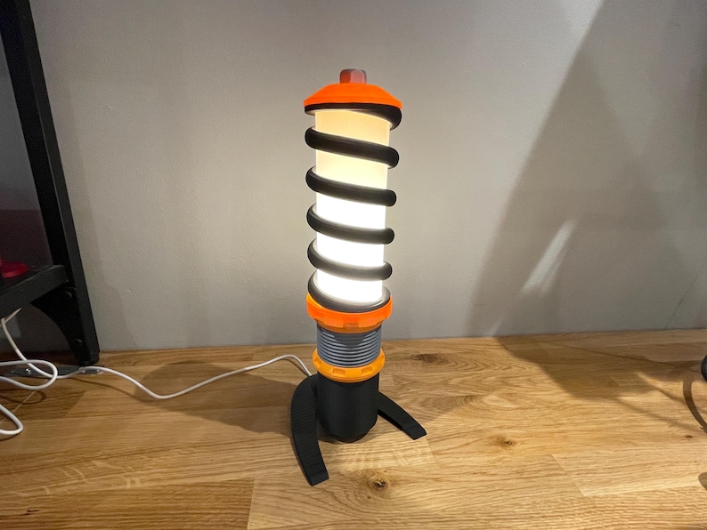 Op de afbeelding: Een zwarte en oranje tafellamp in de vorm van een schokdemper met een witte gloeilamp.