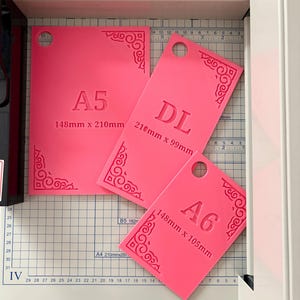 May include: Three bright pink rectangular templates with rounded corner cutouts and decorative scrollwork patterns. Each template is labeled with its size: A5 (148mm x 210mm), DL (210mm x 99mm), and A6 (148mm x 105mm), each featuring a circular hole for hanging.