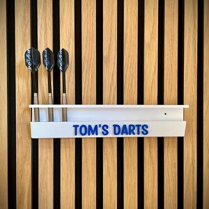 May include: A white dart holder with the text "TOM'S DARTS" in blue lettering, mounted on a wooden wall. Three darts are placed in the holder. The wall has vertical wooden slats in alternating light and dark colors.