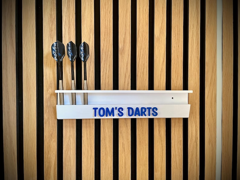 May include: A white dart holder with the text "TOM'S DARTS" in blue lettering, mounted on a wooden wall. Three darts are placed in the holder. The wall has vertical wooden slats in alternating light and dark colors.
