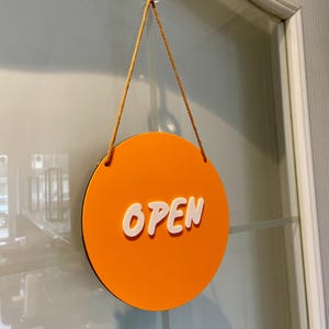 May include: Orange round sign with the word "OPEN" in white lettering. The sign is hanging from a glass door with a suction cup and string.