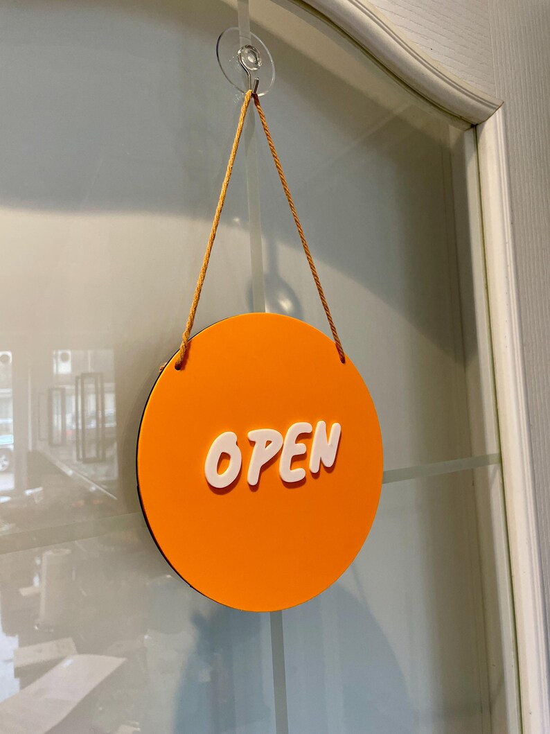 May include: Orange round sign with the word "OPEN" in white lettering. The sign is hanging from a glass door with a suction cup and string.