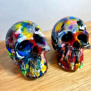 May include: Two colourful, hand-painted human skulls. The skulls are painted in a variety of bright colours, including red, yellow, blue, green, and purple. The skulls are made of a smooth, white material and have a glossy finish.