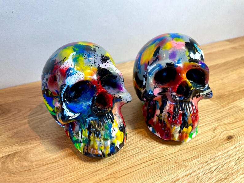 May include: Two colourful, hand-painted human skulls. The skulls are painted in a variety of bright colours, including red, yellow, blue, green, and purple. The skulls are made of a smooth, white material and have a glossy finish.