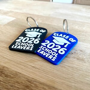 May include: Two graduation-themed keychains. One is black, the other blue. Both have a white graduation cap graphic and the text "CLASS OF 2026 SCHOOL LEAVERS". Each keychain has a silver metal ring attached.