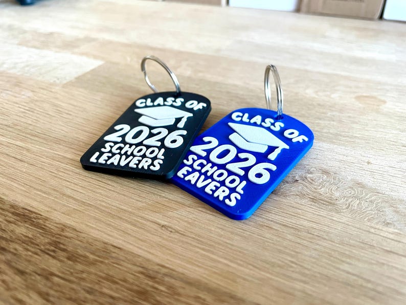 May include: Two graduation-themed keychains. One is black, the other blue. Both have a white graduation cap graphic and the text "CLASS OF 2026 SCHOOL LEAVERS". Each keychain has a silver metal ring attached.