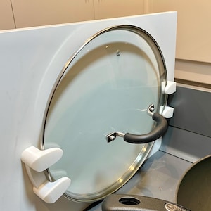 May include: A white cabinet with a glass pot lid attached to the side using white plastic hooks. The lid has a black handle. A black frying pan is in the foreground.
