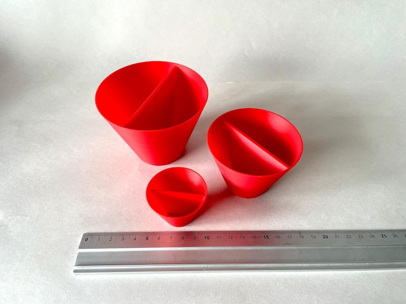 May include: Three red, cone-shaped objects with a central divider are displayed. A metal ruler with inch markings is placed next to the objects. The objects vary in size, with the largest approximately 4 inches in diameter.