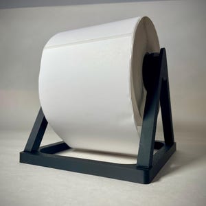 May include: A large roll of white labels sits on a black label holder. The label roll is centered in the frame, with the black holder providing support. The background is a neutral gray.